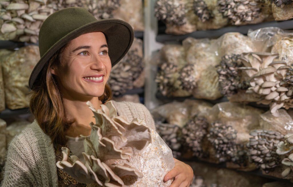 happy young woman smiling and holding grown medium with mushrooms against the oyster mushroom farm. Copy space